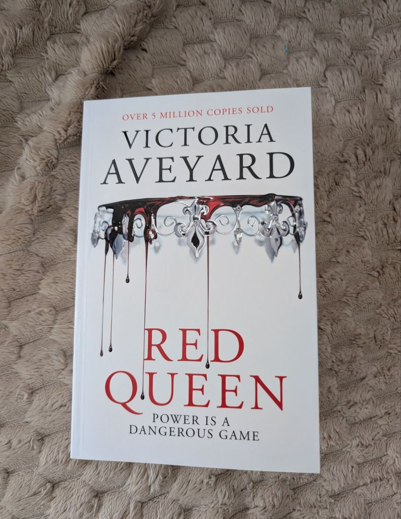 This is the cover of Victoria Aveyard's book, Red Queen. The cover is white with an upside down silver crown and red liquid dripping from it.